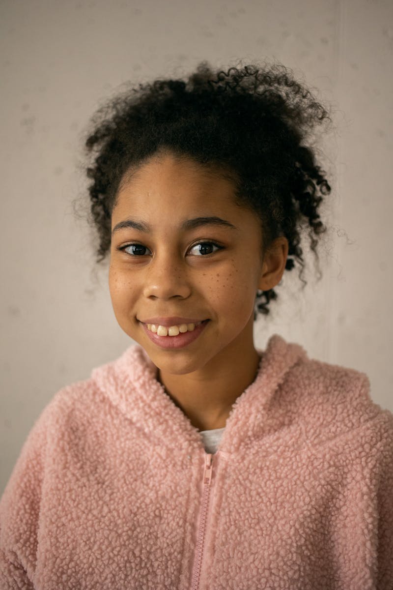 A cheerful young girl with curly hair smiling warmly in a cozy pink sweater indoors.