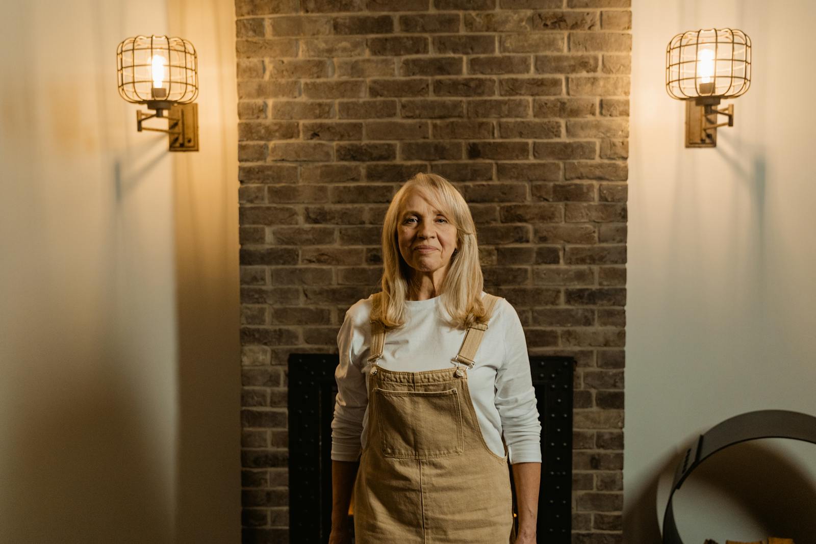 Warm indoor portrait of a senior woman with long hair, standing against a brick wall.