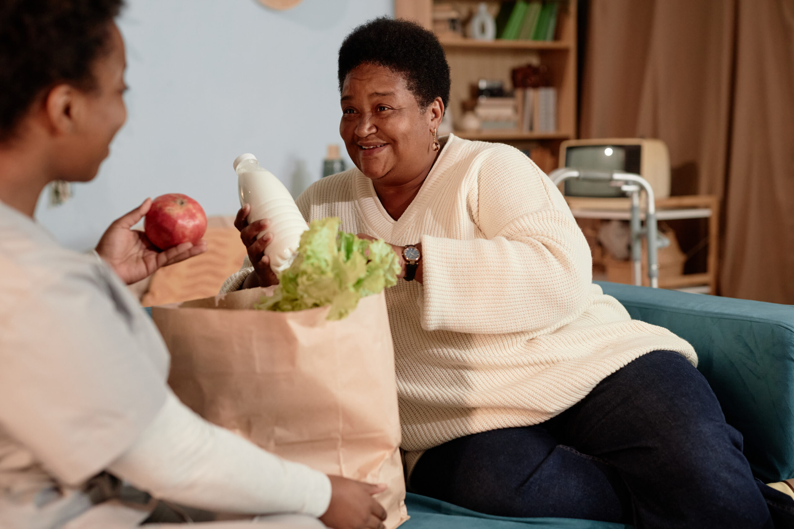Smiling Black Senior Woman Unpacking Bag with Groceries Receiving Help from Social Worker at Home