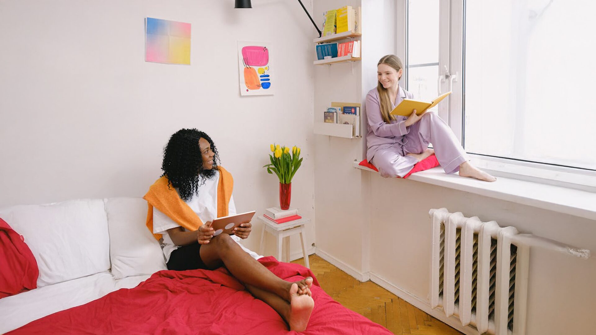 Two women enjoying a cozy moment in a bright, colorful bedroom with books and a tablet.